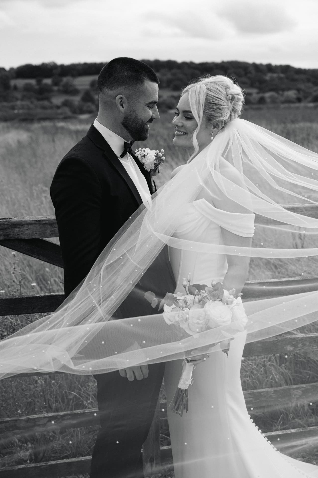 Black and white portrait of a bride and groom by a wooden fence with her veil drifting across the frame